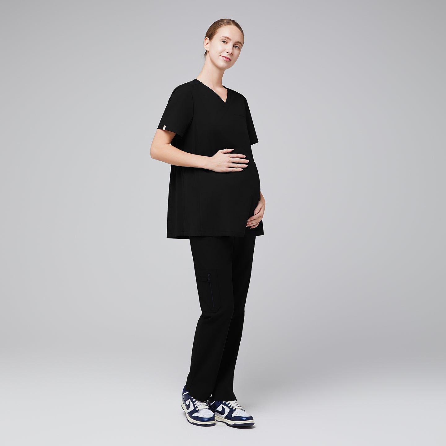 Image of Model wearing black maternity scrub top and matching scrub pants, with her hand holding her pregnancy bump.